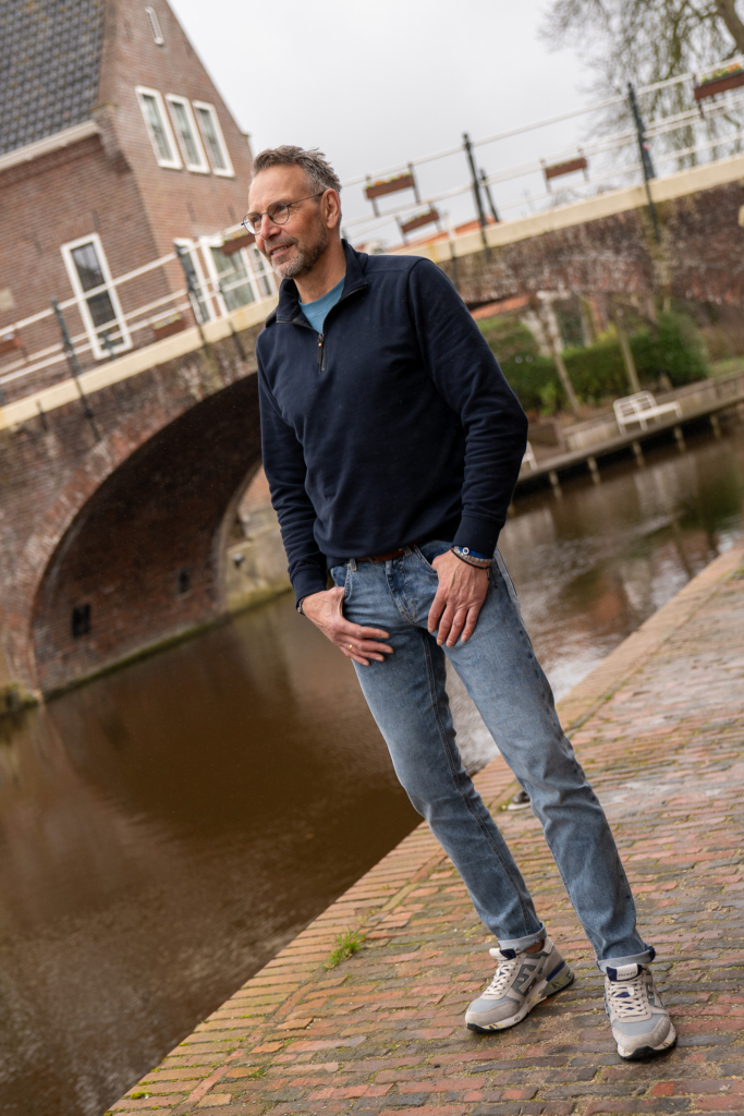 stoere volwassen man, uitkijken over water in Winsum, mooiste dorp van Nederland, spijkerbroek MUD jeans, duurzame jeans, sustainable fashion, sweater Ma-Strum in donkerblauw, verkrijgbaar bij Sytha
