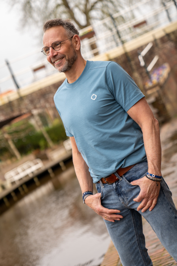 lichtblauw t-shirt Ma-strum gedragen door man van middelbare leeftijd, kijkt blij, staat bij water in Winsum, duurzame mode
