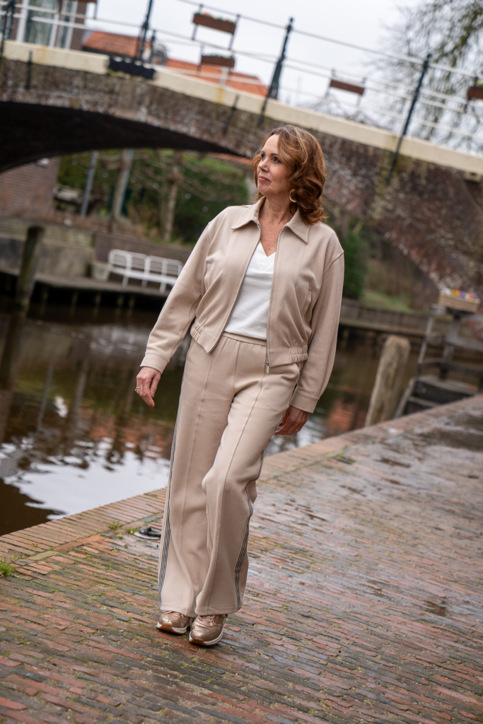 Aimeé the Label luxe trainingspak in créme. Broek met sportieve zwart/witte streep, sporty fashion, créme niet doorschijnend t-shirt, vrouw loopt bij water
