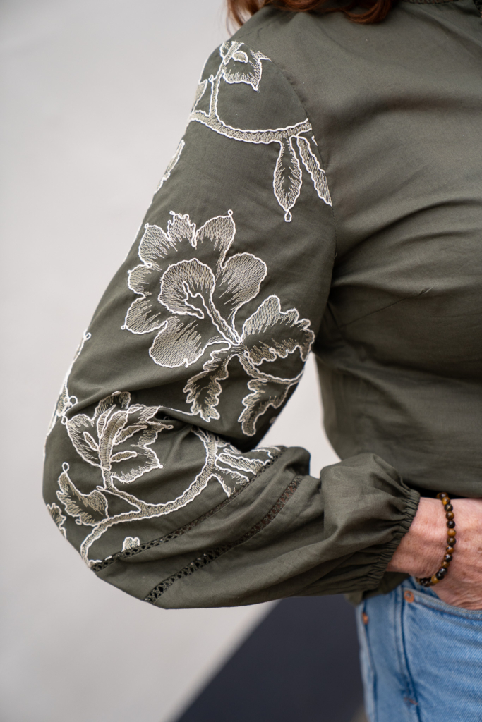 detailfoto van creme borduursel bloemen op rechtermouw van donkergroene blouse met knoopjes voor dames.