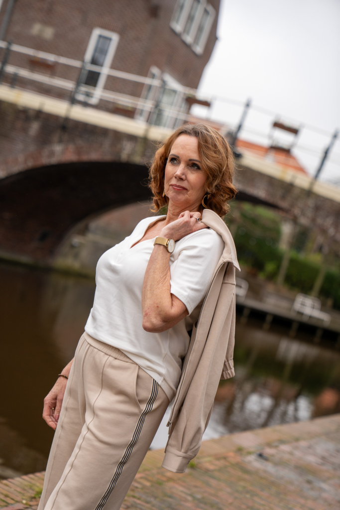 vrouw uitkijkend over water in sportieve set van Aimeé the Label, vlakbij bekende brug in Winsum, vestje créme draagt ze over schouder, comfortable damesmode