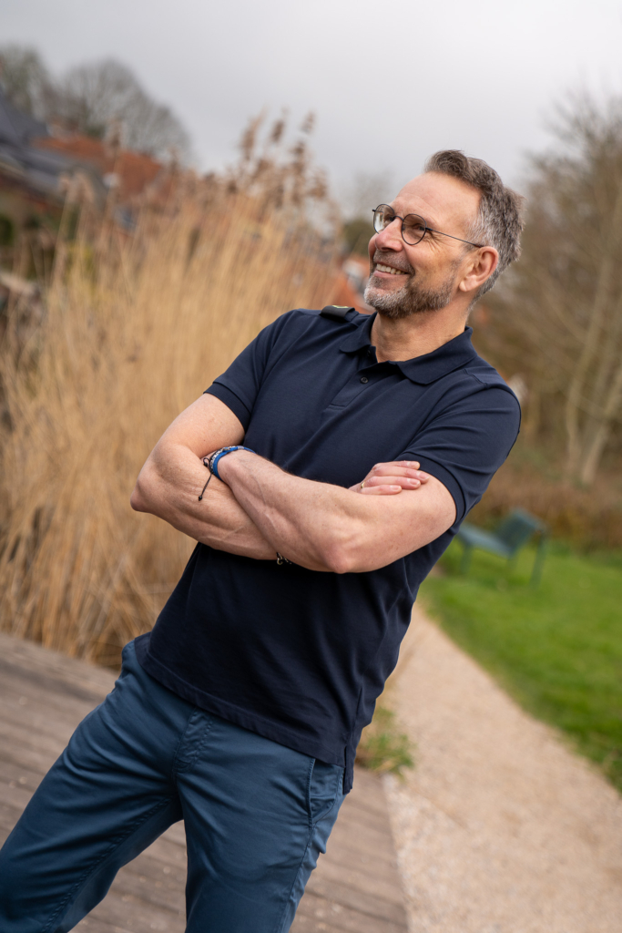 stoer kijkende man met armen over elkaar gekleed in een donkerblauwe navy herenpolo in maat L van Ma-Strum, broek van MUD jeans, duurzaam winkelen Winsum