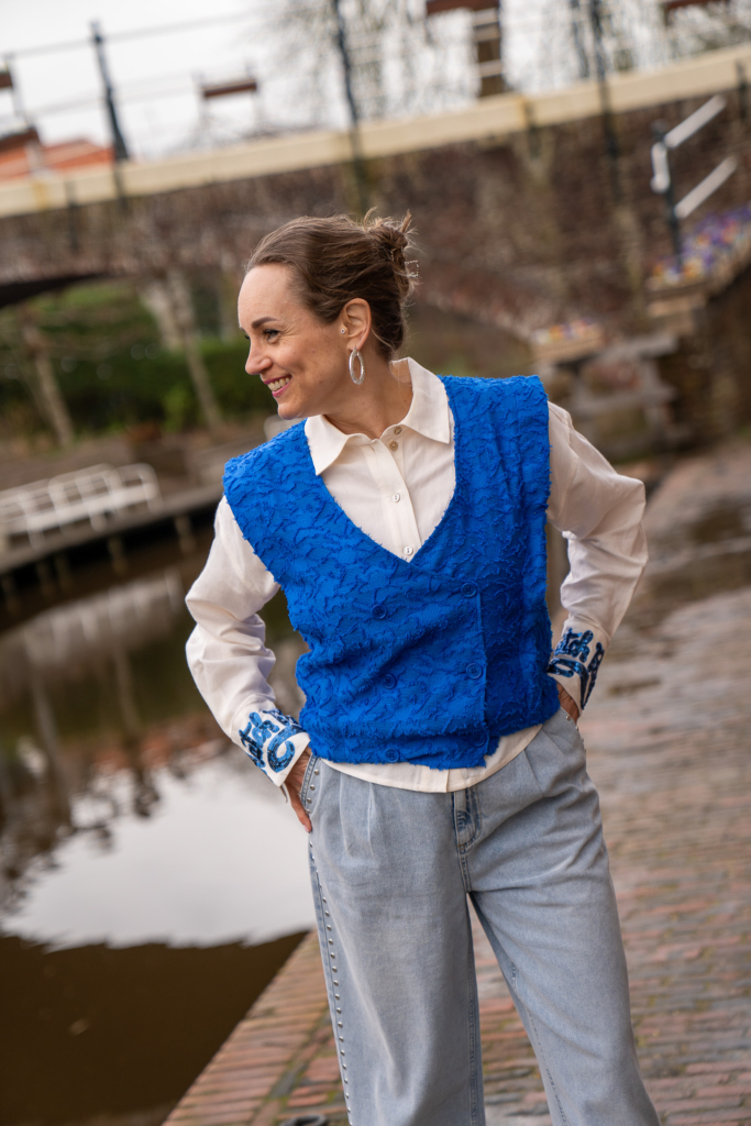 vrouw lacht en kijkt over het water in Winsum, ze draagt een creme linnen blouse met een koningsblauw gilet/spencer van POM Amsterdam