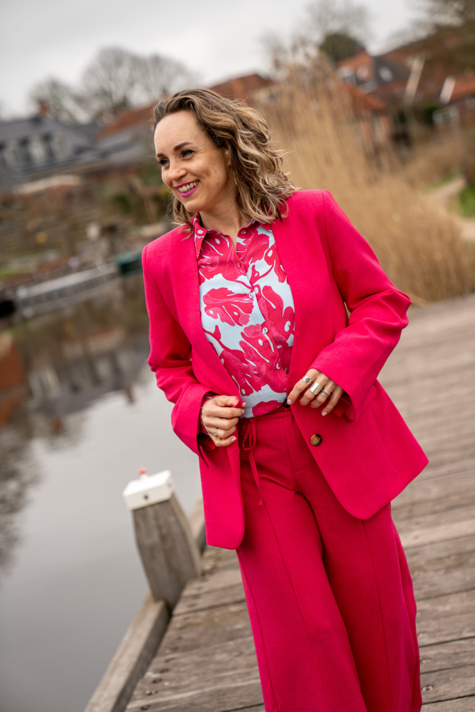 lachende vrouw in fuchsia rose pak van POM Amsterdam, daaronder draagt ze een lichtblauw met rose geprint blousje.